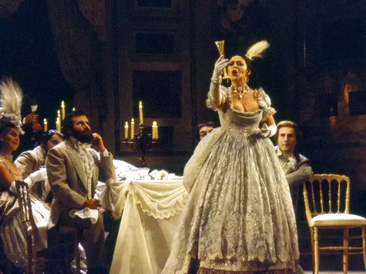 Singer Catherine Malfitano raises a goblet as she sings a drinking song during the opera la Traviata while other performers watch sitting down at a table with white tablecloth