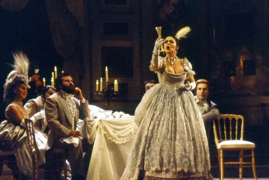 Singer Catherine Malfitano raises a goblet as she sings a drinking song during the opera la Traviata while other performers watch sitting down at a table with white tablecloth