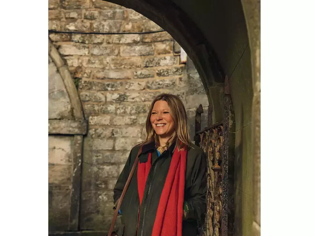 The author Caroline Eden in a red scarf and khaki jacket standing in an arched stone doorway smiling.