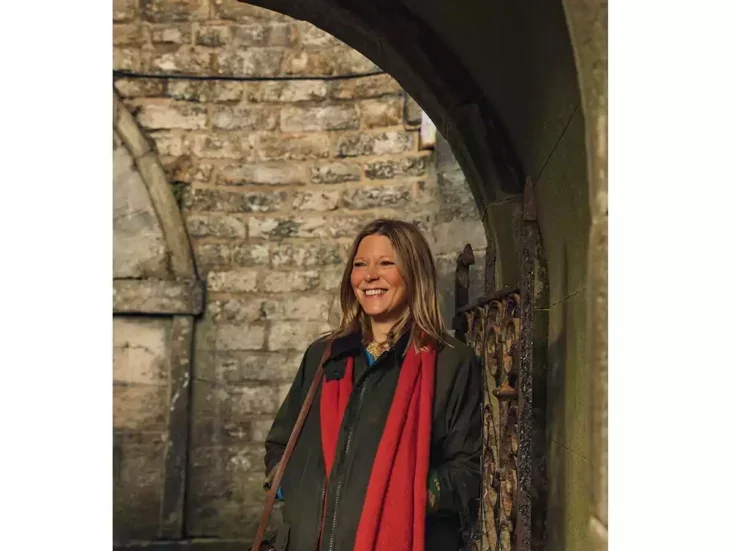 The author Caroline Eden in a red scarf and khaki jacket standing in an arched stone doorway smiling.