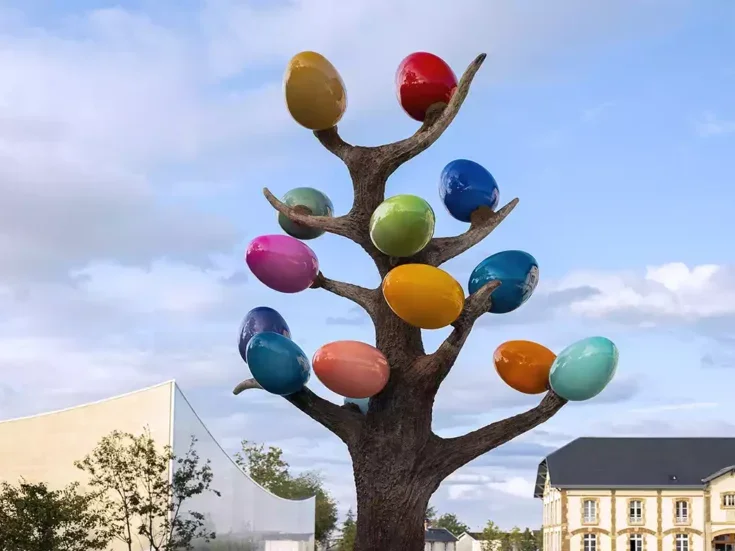 An artwork featuring brightly coloured glass balls on a tree-like structure with branches between two white buildings, one modern and one traditional with a slate grey roof at Ruinart in Reims.