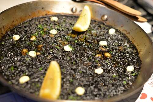 close-up of arroz negro in a metal pan with two lemon wedges and flecks of parsley and aioli.