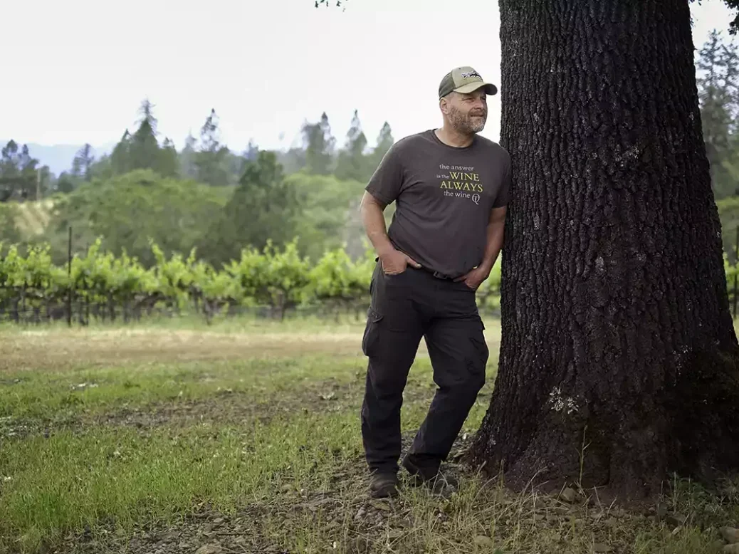 Mike Dunn in baseball cap and t-shirt leans on a tree in Howell Mountain against a backdrop of woodland and vineyard.