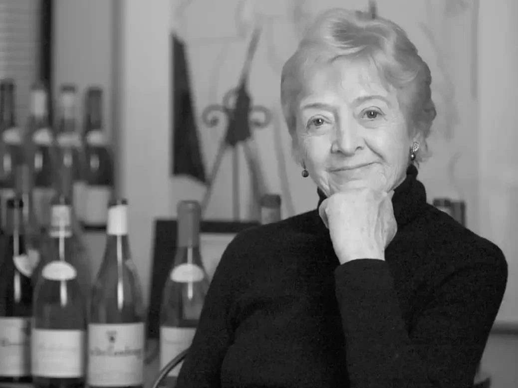 A black and white image of Martine Saunier in a black polo neck sitting in front of a shelf of wines with her hand under her chin