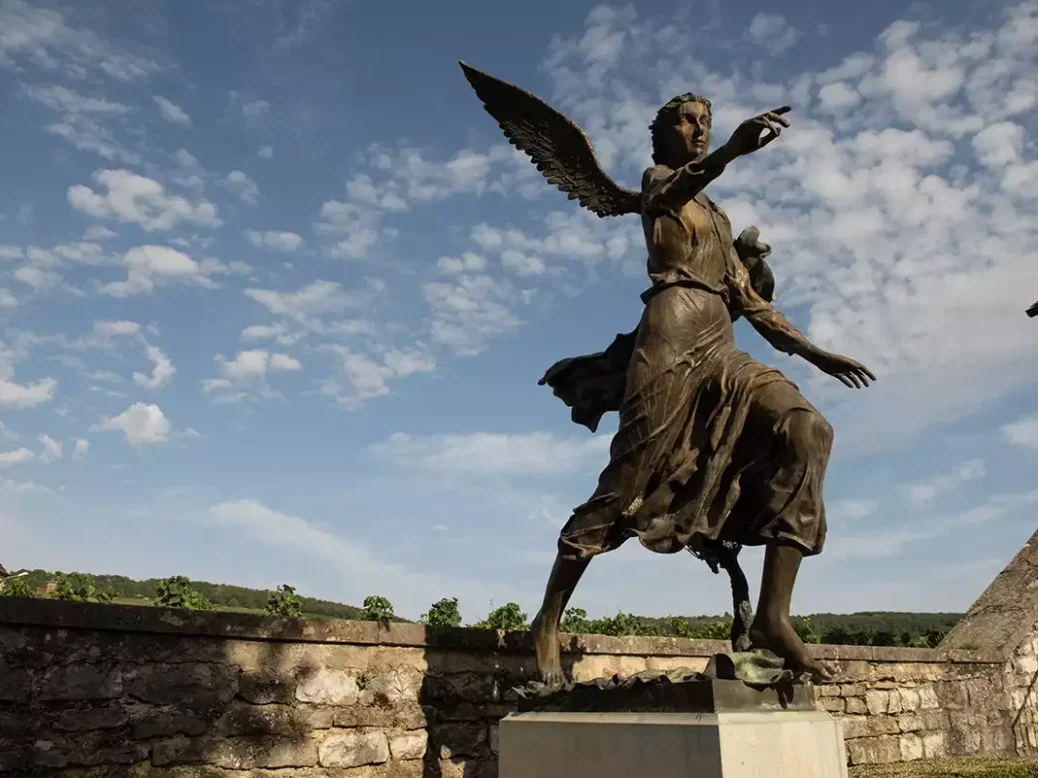 A metal statue of angel pointing against a blue sky with little clouds at Domaine de la Romanée-Conti