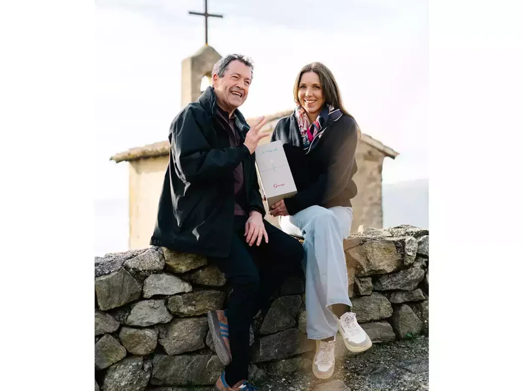 Peter Gago dreessed in black and Caroline Frey both sitting on a stone wall holding a bottle of Grange La Chapelle in front of a chapel with a cross on top