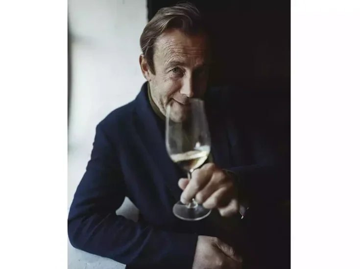 Olivier Collin dreessed in black and leaning on a window sill holding a glass of his Champagne in front of his chin
