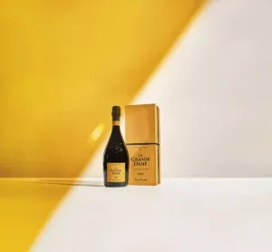 A bottle of 2018 Veuve Clicquot La Grande Dame Champagne beside a yellow box on a white table against a white wall which is half painted along a diagonal in the yellow of Veuve Clicquot