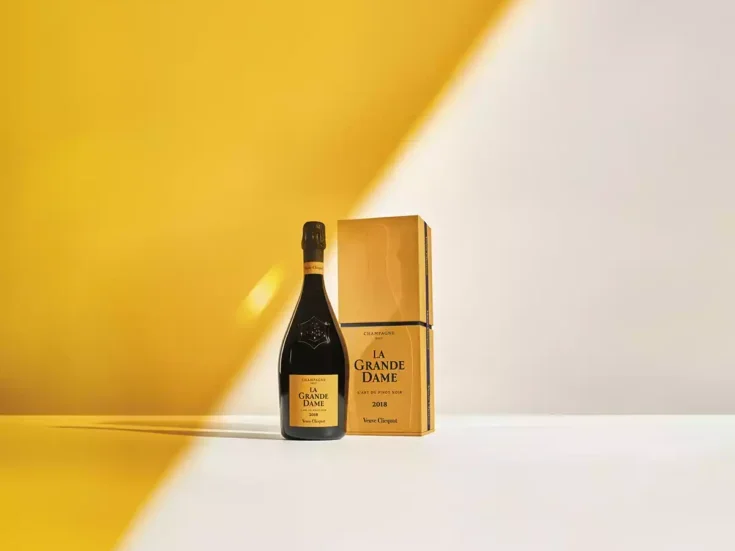 A bottle of 2018 Veuve Clicquot La Grande Dame Champagne beside a yellow box on a white table against a white wall which is half painted along a diagonal in the yellow of Veuve Clicquot