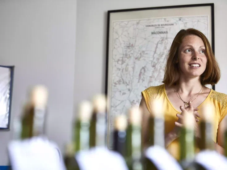 Elodie Rousselot winemaker with red hair and a yellow top standing behind a row of bottles in front of a map