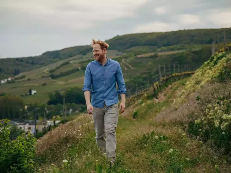 Georg Rumpf, with red hair in a blue shirt and khaki chinos, walking in his vineyards