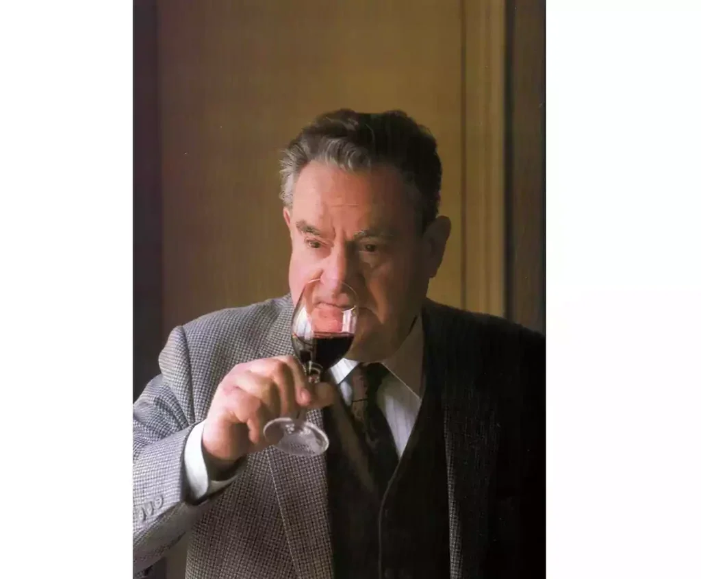 A photo of Emile Peynaud in grey suit and dark tie drinking a glass of red wine