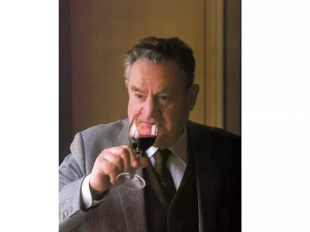 A photo of Emile Peynaud in grey suit and dark tie drinking a glass of red wine
