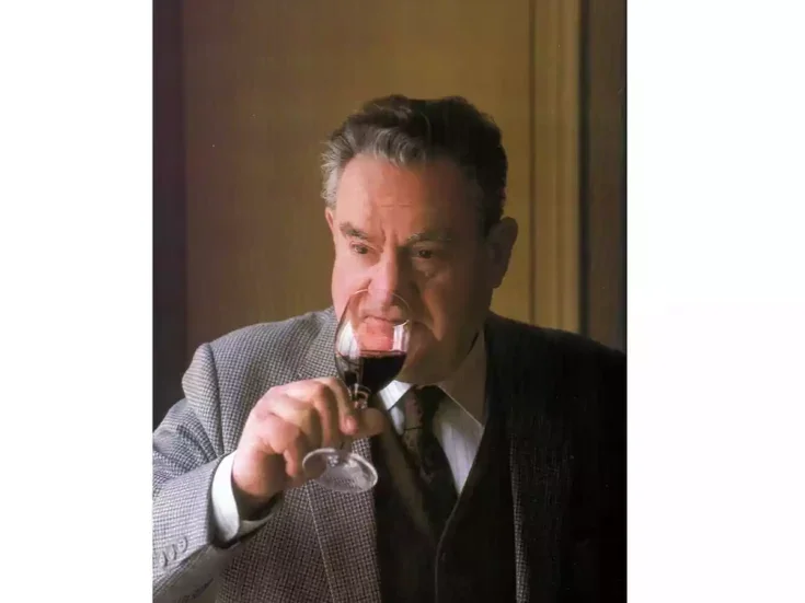 A photo of Emile Peynaud in grey suit and dark tie drinking a glass of red wine