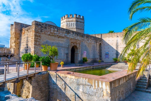 A view of the alcazar / Moorish castle in Jerez de la Frontera with palm trees on the right and a small bridge leading to the stone buildings.