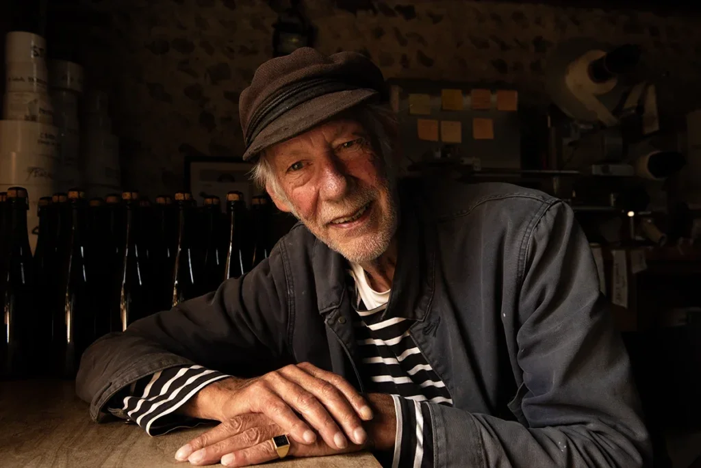 Peter hall with white beard black cap breton shirt and cotton jacket leaning on a table