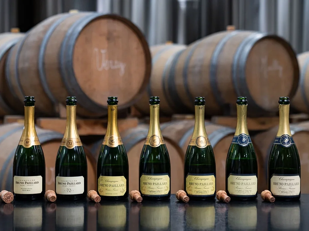 A row of bottles of Bruno Paillard Champagne made using the house's Perpetual Reserve on a table in front of some stacked barrels