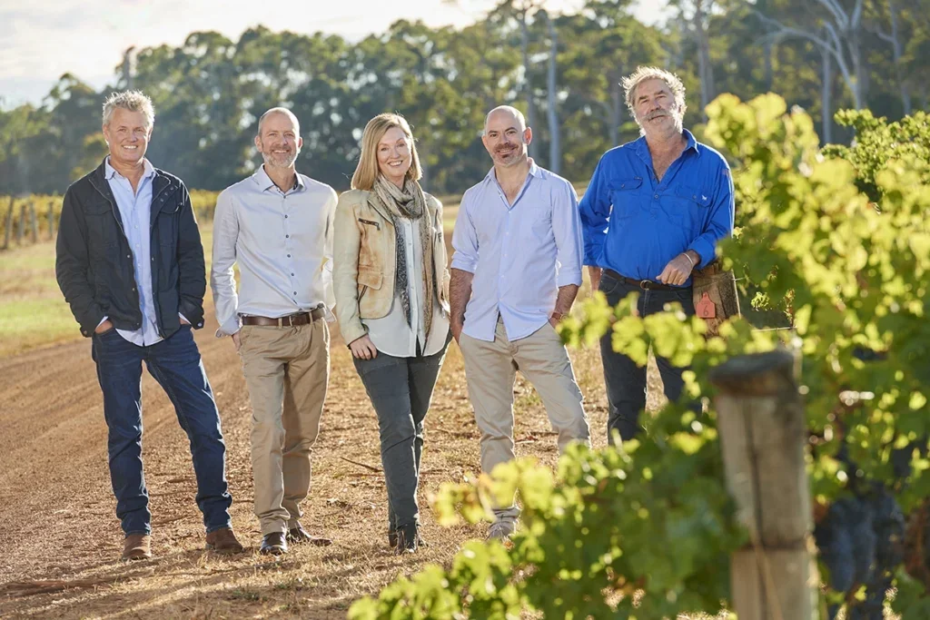 Lined up and smiling for the camera, long blonde-haired Simone Furlong in faded cream jacket flanked by two men on either side on a dusty track through a vineyard