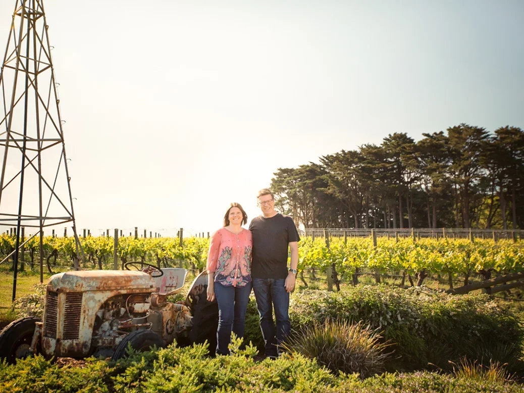 Martin and Karen Spedding stand in their vineyard beneath a lattice-framed metal windmill on a sunny day with trees in the background
