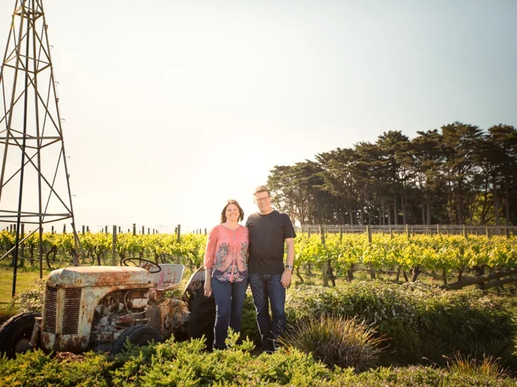 Martin and Karen Spedding stand in their vineyard beneath a lattice-framed metal windmill on a sunny day with trees in the background