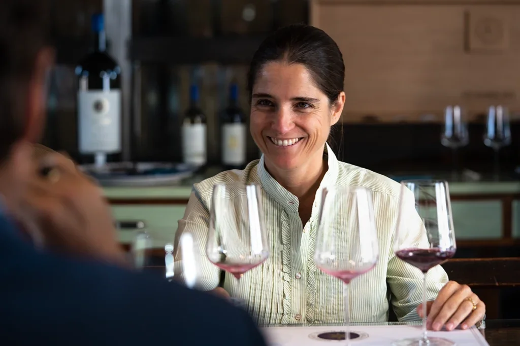 Priscilla Incia della Rocchetta in white stripy shirt with black hair in a pony tail smiling sitting down with three wine glasses, two empty, one with red wine