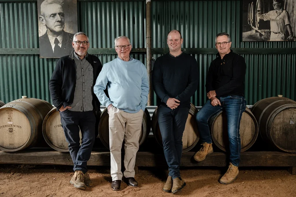 Four men of varying ages standing in a line smiling at the camera in front of a row of barrels in a corrugated metal barn
