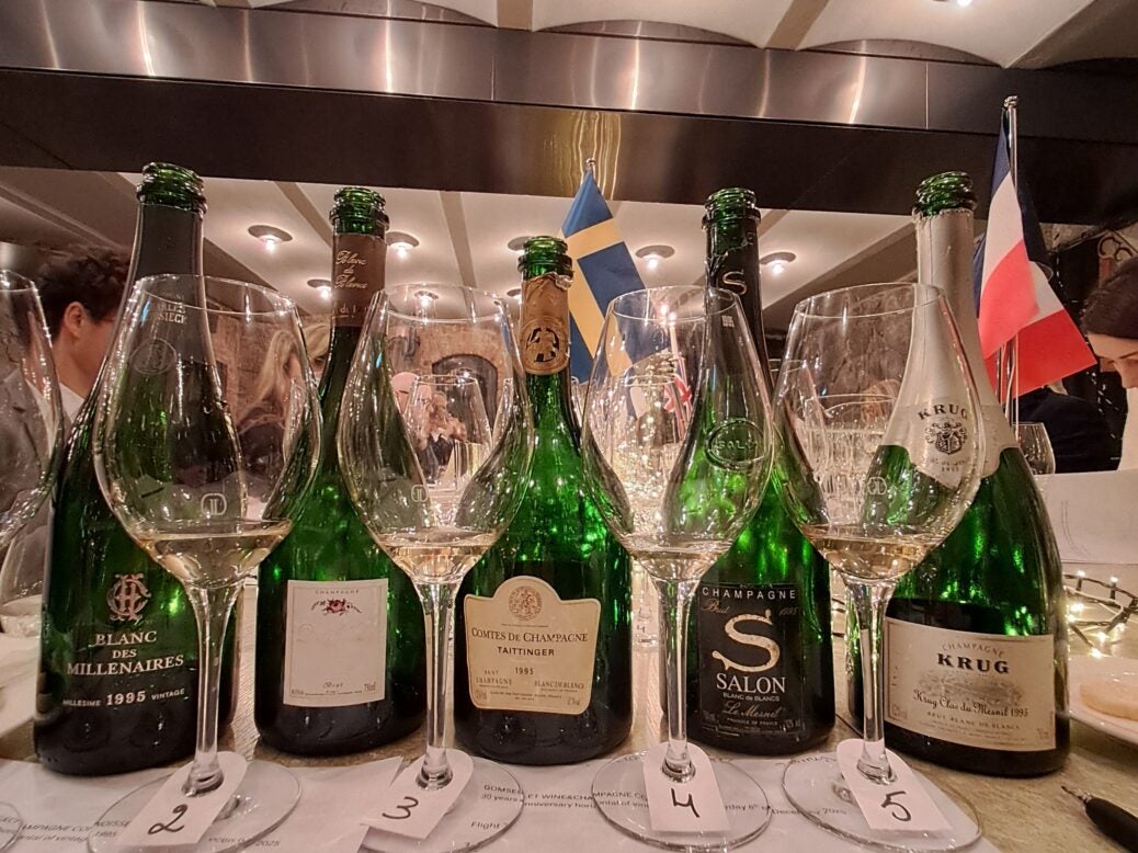 Five bottles of 1995 Vintage Champagne behind four Champagne glasses on a white table cloth in a restaurant with lighs in the ceiling