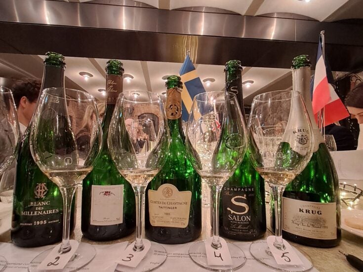 Five bottles of 1995 Vintage Champagne behind four Champagne glasses on a white table cloth in a restaurant with lighs in the ceiling