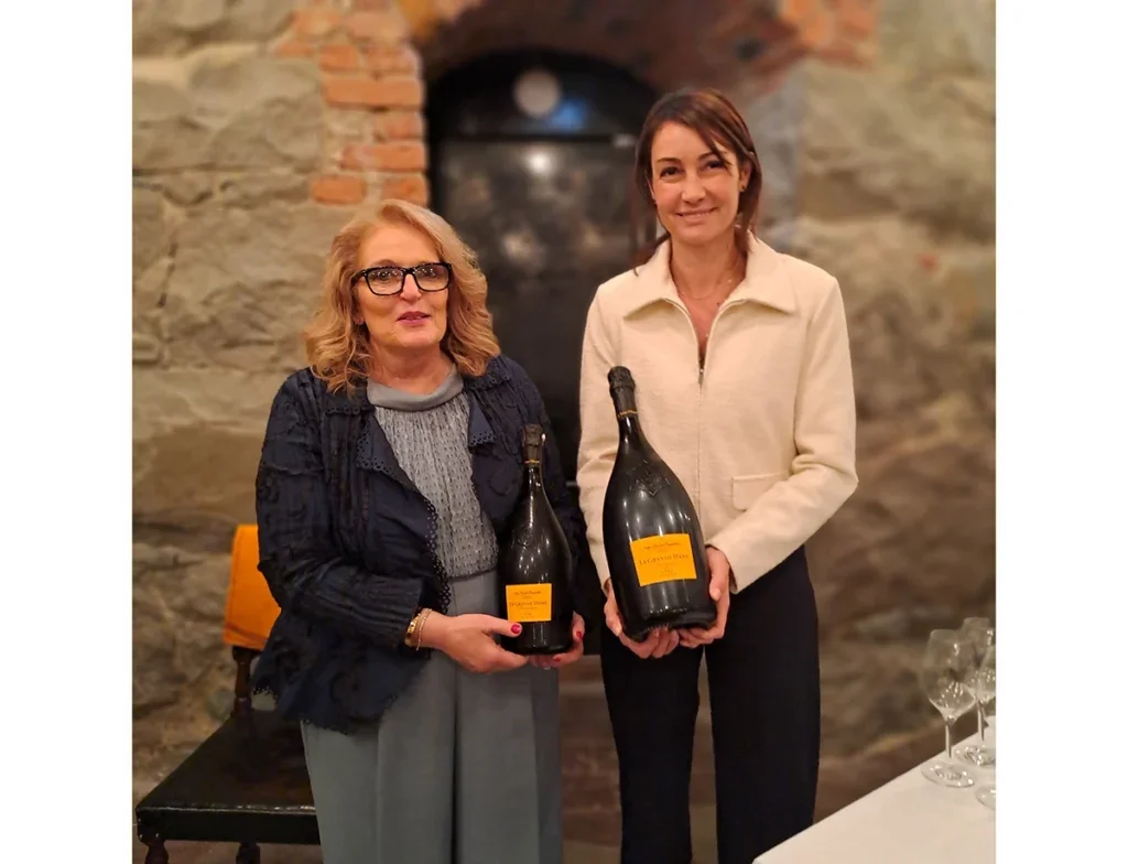 Marina Olsson in glasses and shoulder length blonde wavy hair and smart blue jacket and dress stands next to Gaëlle Goossens with short brown hair in white shirt and black trousers holding two bottles of Champagne