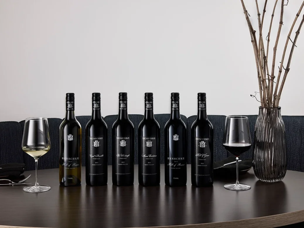 A row of black-labelled Henschke bottles on a shiny black surface with a vase of twigs against a white background