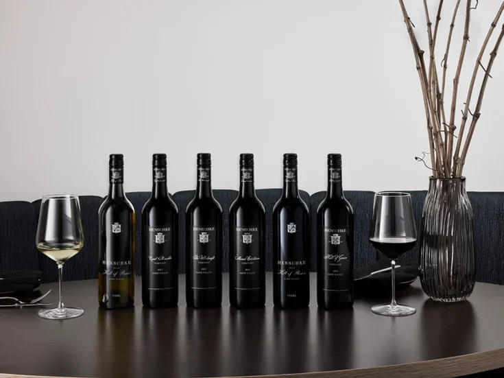 A row of black-labelled Henschke bottles on a shiny black surface with a vase of twigs against a white background