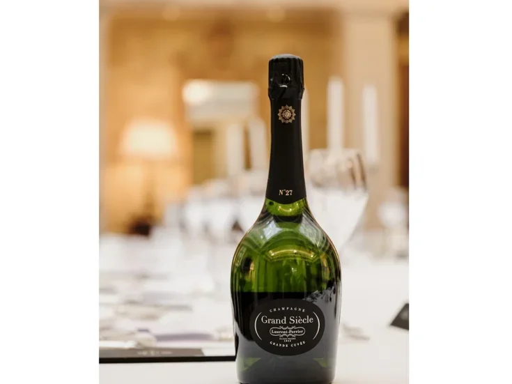 A bottle of Laurent-Perrier Grand Siècle on a white table cloth with some glasses and cutlery in a brightly lit restaurant.