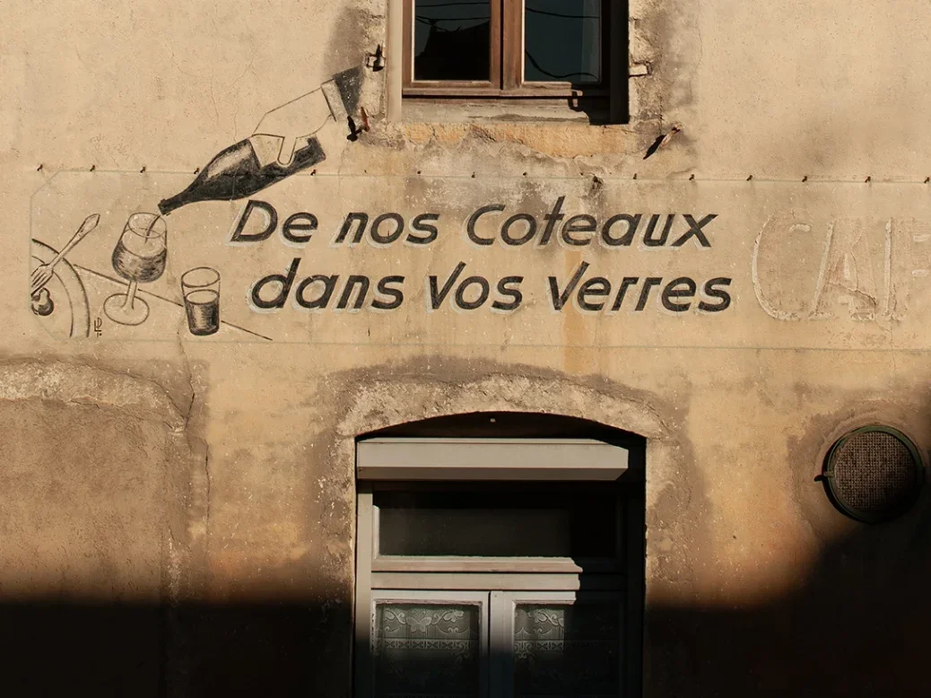 An illustration painted in black and white of a bottle of wine pouring into three glasses on a sandstone wall of an old house in Burgundy, the bottle looks like it's coming out of the window.