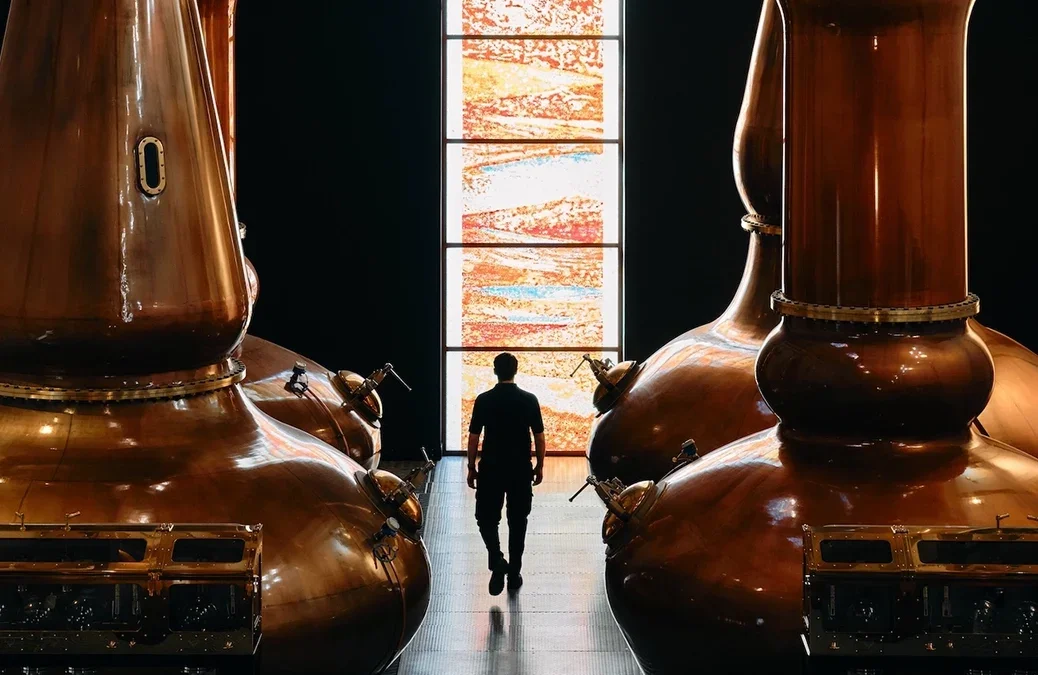 A man walking between two enormous copper stills towards a strip of stained glass window at The Dalmore distillery, a stained gla
