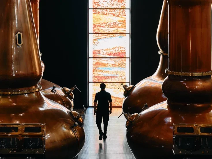 A man walking between two enormous copper stills towards a strip of stained glass window at The Dalmore distillery, a stained gla