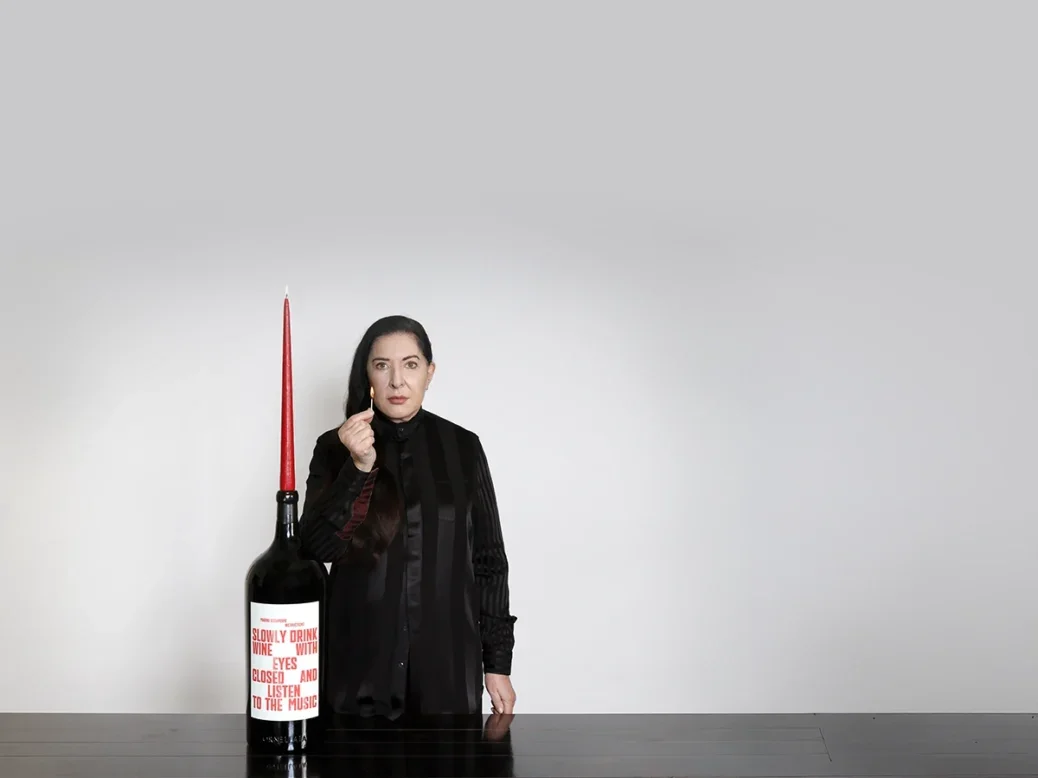 Marina Abramovic in black dress standing with impassive gaze next to a large bottle with red writing on a while label and a long, thin, lit red candle coming out of the top. Abarmovic is holding a lighted match