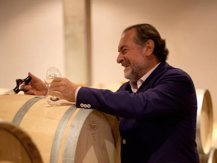 Michel Rolland in dark suit happily looking at a barrel with a large pipette