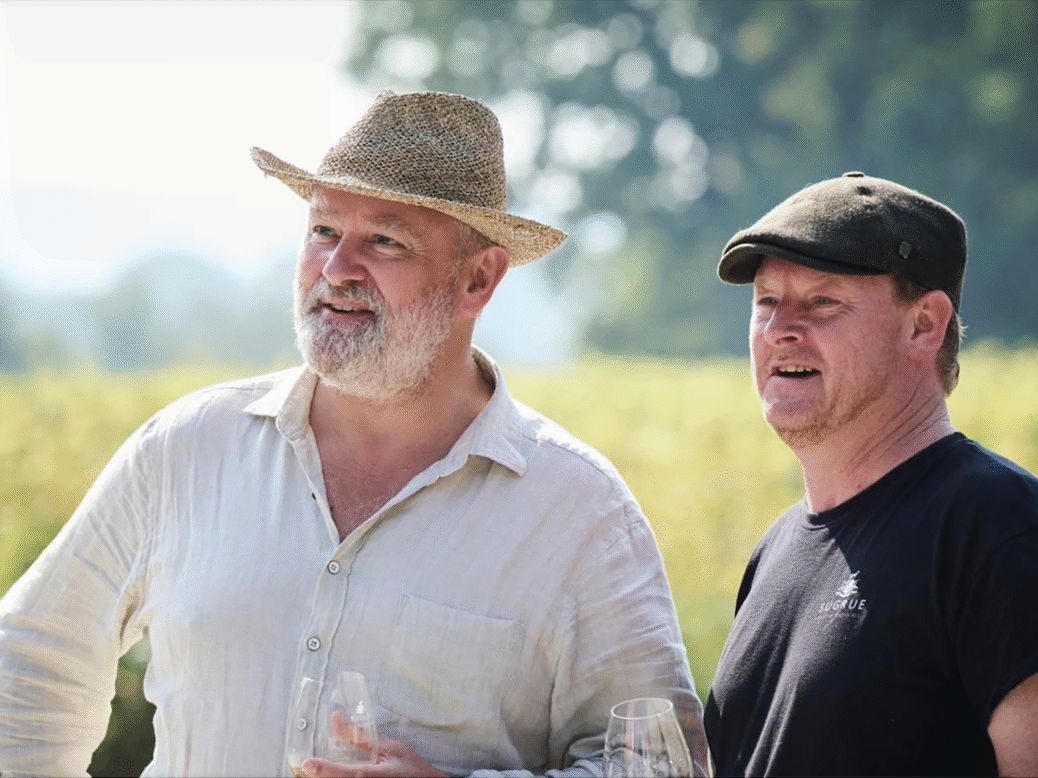 Hugh Bonneville in white shirt, white beard and straw hat stands next to Dermot Sugrue in black t-shirt and black flat cap on a warm summer's day by a vineyard