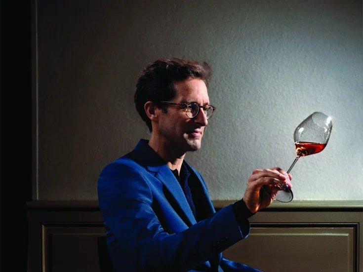 Vincent Chaperon in blue jacket and t-shirt, glasses and curly hair looking at a glass held in front him at an angle