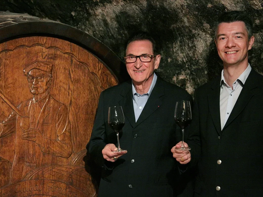 Philippe Guigal (right) and Marcel both in black jackets and open-necked shirts holding glasses of wine by a big barrel with a relief of a winemaker
