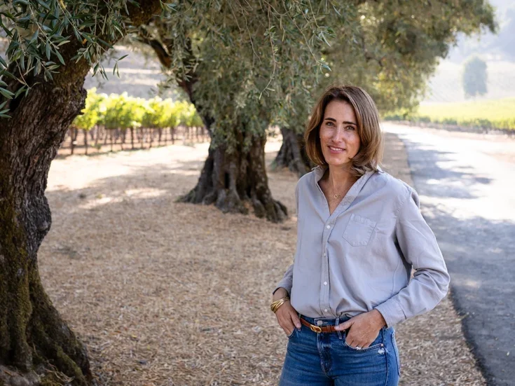 Kathryn Green in blue tucked in shirt and jeans and shoulder length brown hair standing on a tree-lined path beside her vineyard.