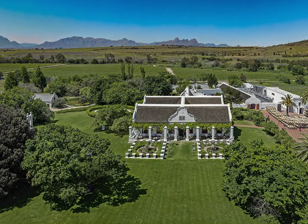 Aerial view of Meerlust estate, a white Cape Dutch house surrounded by green and mountains in the distance on a bright blue-sky day