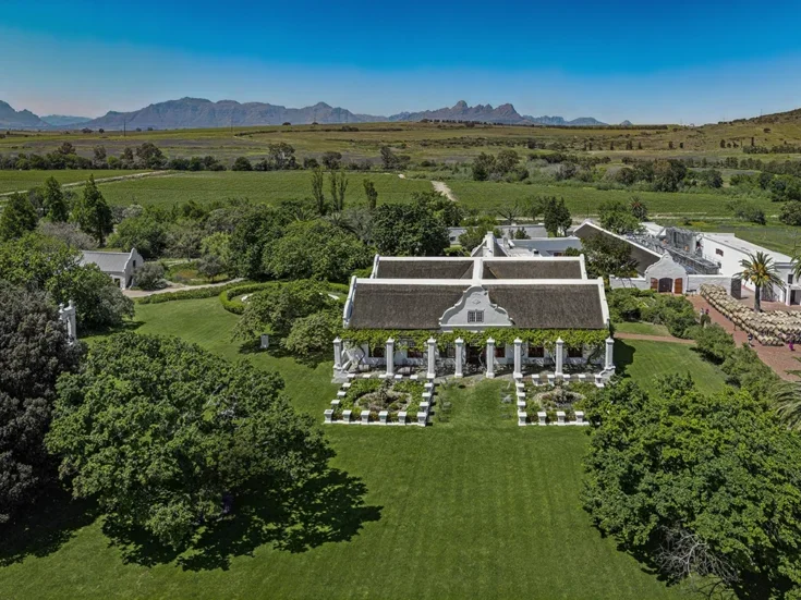Aerial view of Meerlust estate, a white Cape Dutch house surrounded by green and mountains in the distance on a bright blue-sky day
