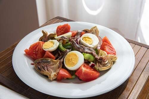 A white plate with a salade niçoise of eggs, achovies, tomatoes on a slatted brown wooden table with a white curtain or table cloth in the sun behind