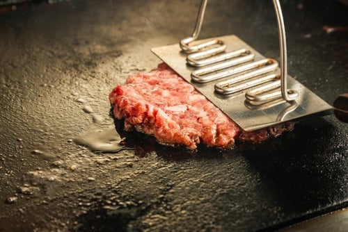 A single raw burger on a flat griddle being pressed down on with a metal device for flattening burgers