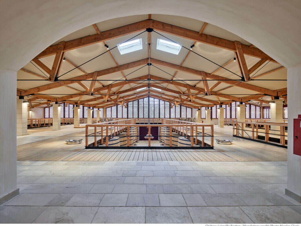 The winery of Léoville Barton with wooden framed white roof and a balcony above large oak barrels. Very clean and new.