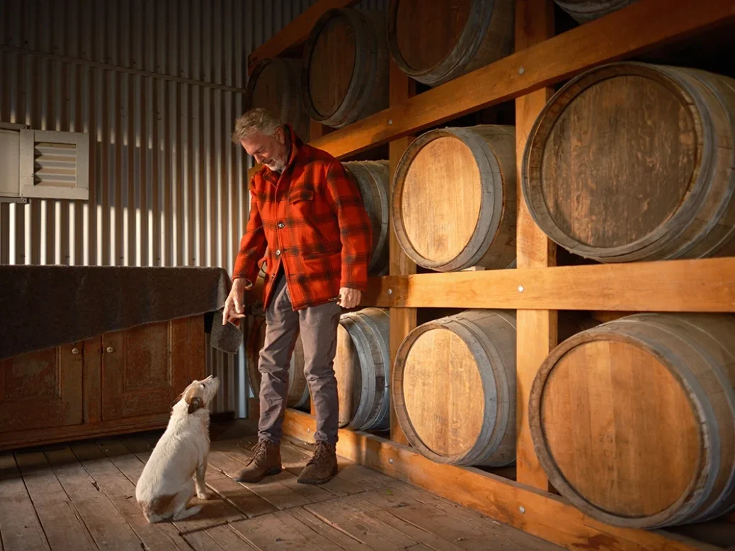 Sam Neill in a cellar with barrels in red and black jacket looking down at a friendly little white dog who looks back at him