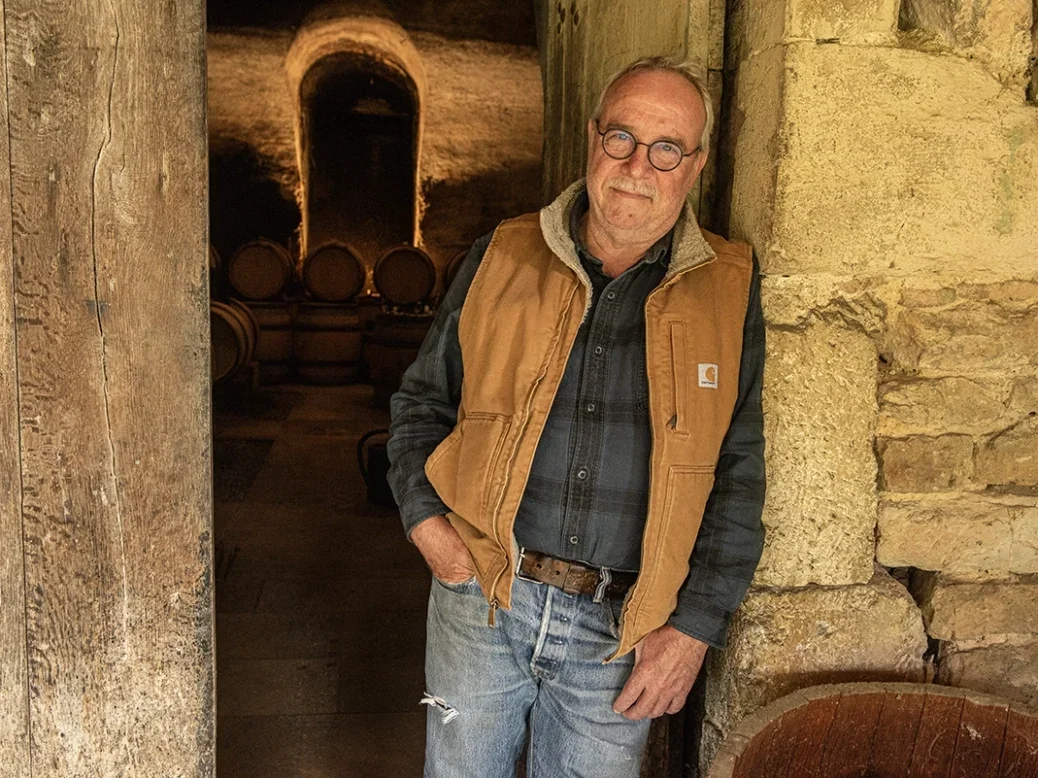Olivier Merlin with white hair and black round framed glasses dressed in jeans, checked shirt and light brown sleeveless fleece, leaning on a stone wall in his cellar