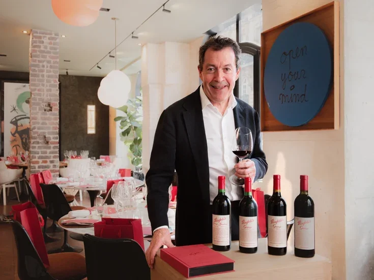 Peter Gago in black suit and white shirt and black hair happy with some bottles of Penfolds wine in a room