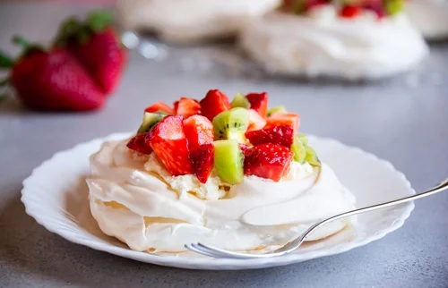 A pavolova topped with strawberries and kiwi fruit on a white plate on a gray table with other pavlovas in the distance